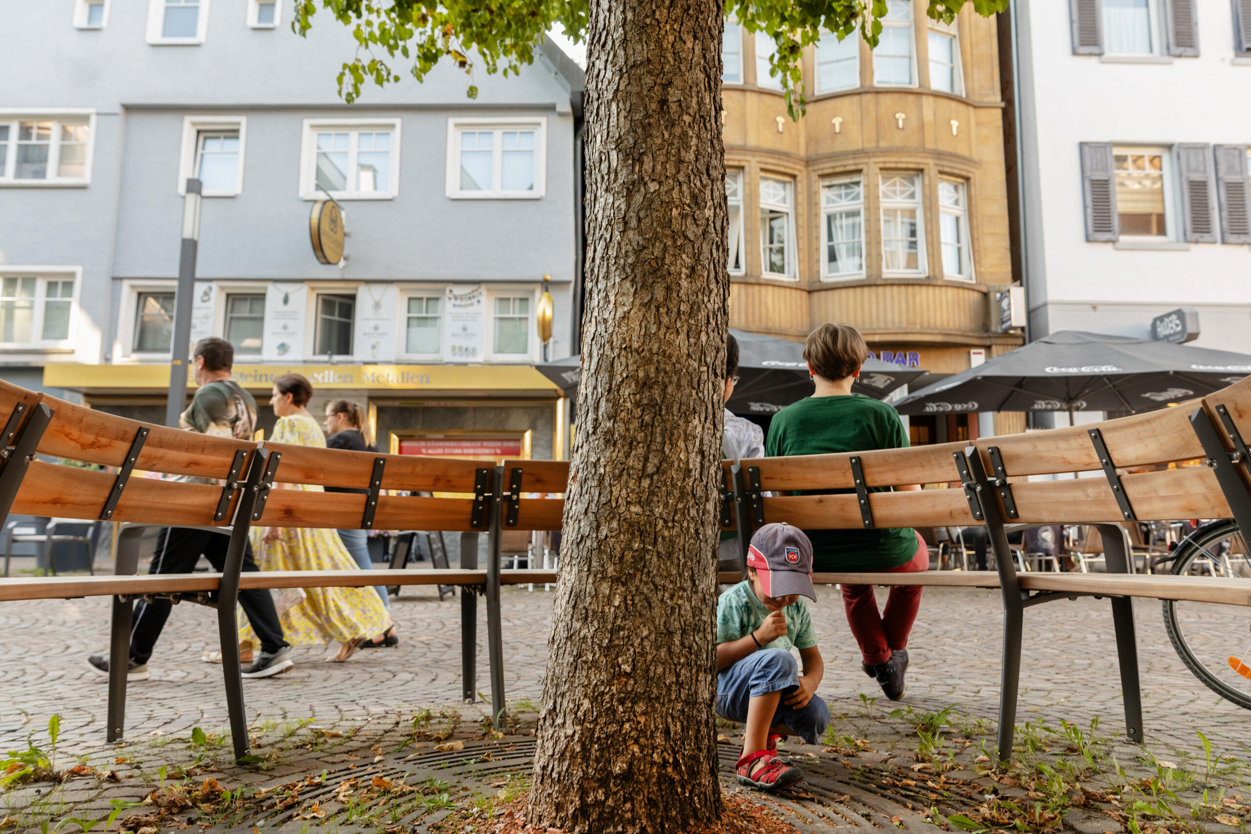 Fotografie einer Rundsitzbank in der Fußgängerzone, im Vordergrund ein Kind an einem Baum spielend